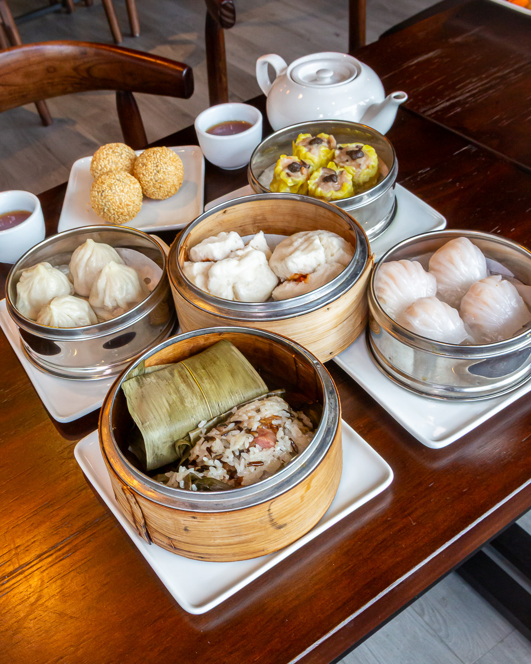 Jo Jo Wok closeup dim sum2849 "A full spread of dim sum dishes at Jo Jo Wok, including assorted bamboo steamers with dumplings, pork buns, sticky rice parcels, a plate of sesame balls, teacups, and a teapot on a wooden table
